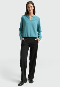 Turkusowy sweter z dekoltem w serek i prążkowanymi detalami, w połączeniu z czarnymi, długimi, prostymi spodniami oraz czarnymi butami. Gładka faktura na całej powierzchni.