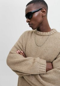 Beige gestrickter Pullover mit einem gerippten Rundhalsausschnitt und oversized Ärmeln. Begleitet von einer schwarzen Sonnenbrille und einer silbernen Kettenhalskette.