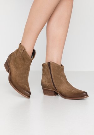 Jambes portant des bottines en daim marron avec des fermetures éclair latérales et des talons blocs bas sur une surface blanche.