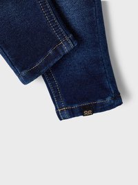 Vaqueros de mezclilla en azul oscuro con una textura ligeramente deslavada, costuras visibles en un dorado en contraste y una pequeña etiqueta negra en el dobladillo.