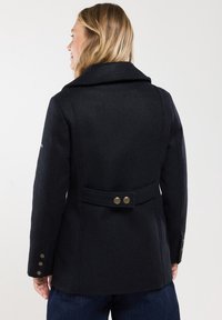 Abrigo de lana negro con un amplio collar, cinturón a la cintura con doble botón y botones metálicos en los puños. Presenta un ajuste entallado y una textura suave.