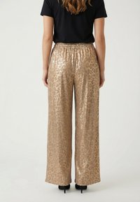 Mujer que lleva pantalones anchos con estampado de leopardo metálico dorado y cintura elástica, combinados con una camiseta negra de manga corta y tacones.