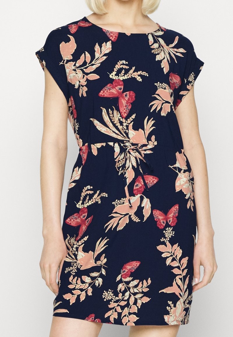 Femme portant une robe bleu marine à manches courtes avec des motifs floraux roses et papillons rouges, avec un détail de ceinture à la taille.