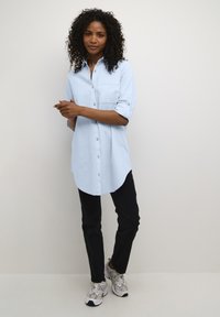 Vaaleansininen nappipaita rintataskulla, käännetyt hihat ja pyöreä helma, yhdistetty mustiin housuihin ja kuvioituihin tennareihin.