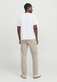 Tricou alb cu mânecă scurtă, croi lejer, purtat cu pantaloni bej deschis și încălțăminte albă cu talpă bej. Fundalul este gri deschis uniform.