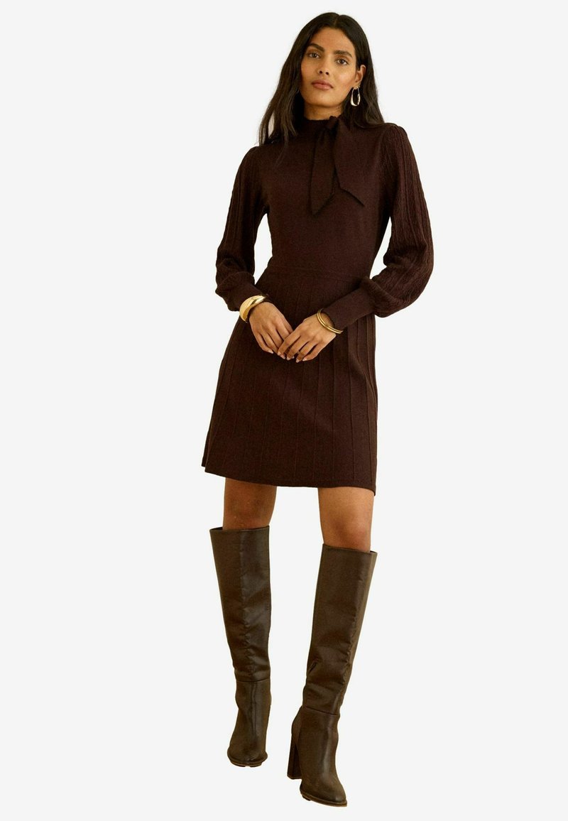 Kobieta ubrana w ciemnobrązową dzianinową sukienkę z długimi rękawami, zapięcie przy szyi, złote bransoletki i ciemnobrązowe, sięgające do kolan botki na obcasie.