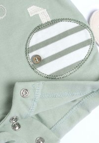 Un body en coton vert avec une patch rayé et un bouton, des fermetures à pression, et des détails de couture contrastés. Texture douce et design simple.