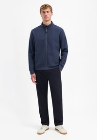 Jonge man die staat, gekleed in een marineblauwe zip-up jas, donkere broek en witte sneakers, tegen een effen witte achtergrond.