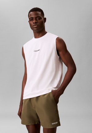 Mężczyzna w białej bezrękawnikowej koszulce Calvin Klein i oliwkowo zielonych szortach Calvin Klein, stoi z jedną ręką w kieszeni na szarym tle.