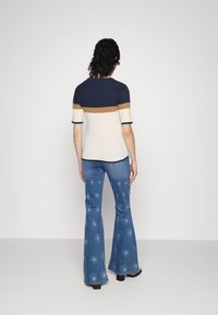 Vrouw die wegkijkt, gekleed in een marineblauwe, beige en bruine korte mouwen trui en een blauwe flared jeans met witte bloempatronen.