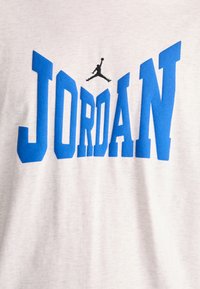 Svijetlosivi pamučni majica s istaknutim plavim slovima koja ispisuju "JORDAN" i malim crnim siluetom figure koja skače iznad teksta.