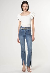 Witte off-the-shoulder top met gerimpelde mouwen, gecombineerd met lichtblauwe denim jeans met vooruitsneden en een gestructureerde zilveren riem.