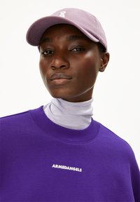 Paarse sweatshirt met een wit logo, gelaagd over een licht lavendelkleurige turtleneck. Een lichtpaarse baseballcap maakt de outfit compleet.
