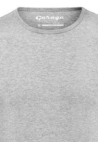 Graues T-Shirt mit kurzem Arm aus weichem Stoff, mit rundem Ausschnitt und einem Etikett mit dem "Garage"-Logo und Größenangabe.