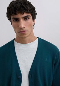 Cardigan teal cu un design în formă de V, două butoane și un logo subtil pe partea stângă, suprapus peste o cămașă gri deschis. Textură netedă.