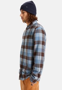 Camicia a maniche lunghe a quadri in tonalità blu e marrone, con apertura anteriore a bottoni, tasca sul petto e una consistenza morbida. Abbinata a un berretto a coste.
