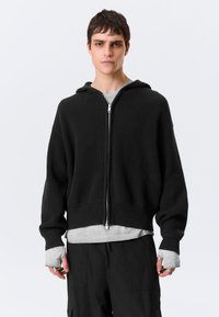 Camisola preta com capuz canelado e fecho de correr, apresenta um corte solto, mangas compridas e capuz com cordão. Usada sobre uma t-shirt cinzenta com gola redonda.