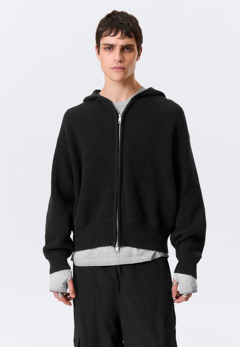 Camisola preta com capuz canelado e fecho de correr, apresenta um corte solto, mangas compridas e capuz com cordão. Usada sobre uma t-shirt cinzenta com gola redonda.