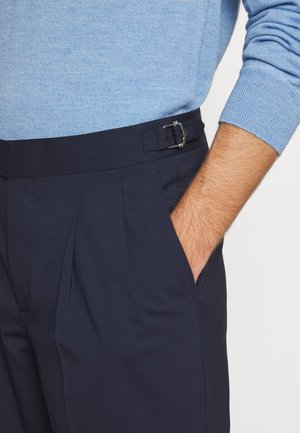 Uomo con maglione azzurro chiaro, mano nella tasca di pantaloni blu navy con dettaglio fibbia laterale in vita.