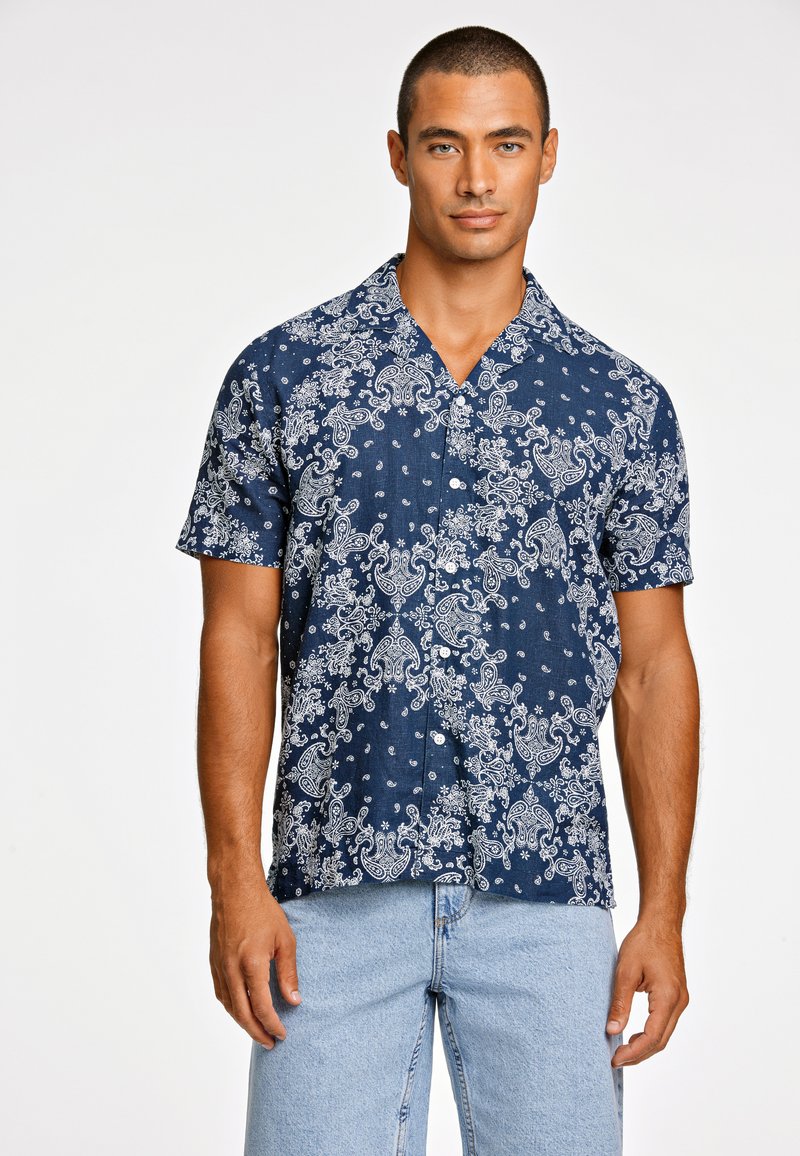 Camisa de manga corta en tejido azul oscuro con un patrón de paisley blanco. Presenta un cuello y cierre de botón. Combinada con pantalones cortos de mezclilla claros.