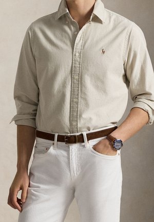 Man draagt een beige overhemd met knopen en opgerolde mouwen, witte spijkerbroek, bruine leren riem en een polshorloge met een blauwe wijzerplaat en een bruine band.