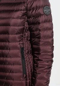 Chaqueta de plumas burdeos con un diseño acolchado, que presenta un bolsillo con cremallera y una etiqueta de parche redonda en el hombro. Textura suave y brillante.