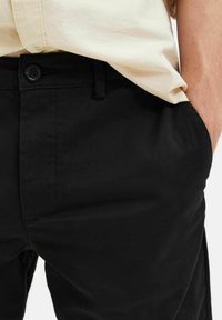Black pants with a smooth texture, featuring a button closure and side pockets, paired with a light-colored shirt.