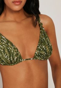 Mujer con un top de bikini triangular verde con estampado y tirantes anudados, mostrado desde el cuello hasta la mitad del torso sobre un fondo neutro.