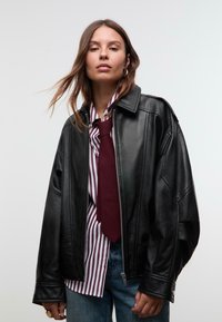 Čierna kožená prešívaná bunda s oversized strihom a zipsom, so zvýrazneným ramenami, doplnená pruhovanou košeľou a vínovou kravatou.