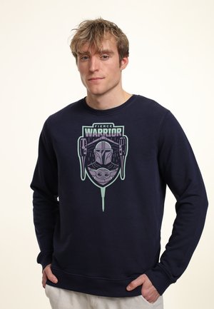 Młody mężczyzna w granatowej bluzie z napisem "Fierce Warrior" oraz grafiką zbrojnego wojownika w hełmie i małego stwora poniżej, stoi z rękami w kieszeniach.