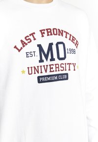 Hvid sweatshirt lavet af bomuld, med rød og marineblå tekst, rund halsudskæring og dekorative stjerner. Teksten siger "LAST FRONTIER UNIVERSITY."