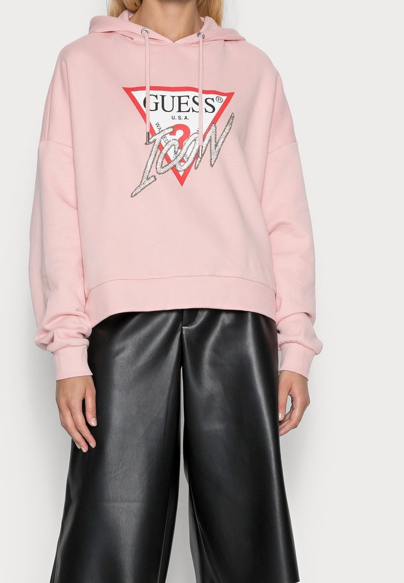 Růžová mikina s červeným trojúhelníkovým logem, na kterém je nápis "GUESS" a "ICON" ve třpytivém písmu. Spárována s širokými kalhotami z černé umělé kůže.