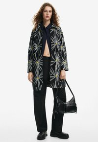 Black coat featuring a floral pattern with gray leaves and yellow centers, paired with a navy shirt and black pants, holding a black handbag.