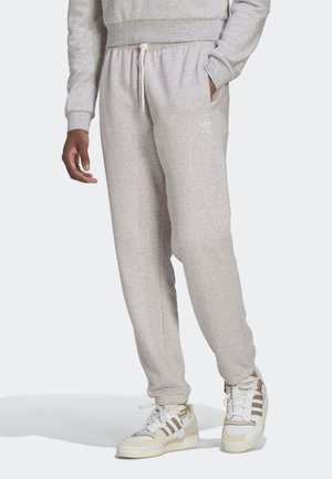 Man in lichtgrijze Adidas-sweatpants en bijpassende sneakers, staand met één hand in zijn zak tegen een effen achtergrond.