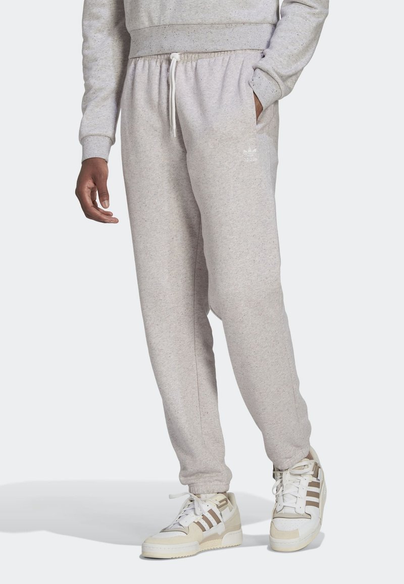 Man som bär ljusgrå Adidas joggingbyxor och matchande sneakers, står med ena handen i fickan mot en enkel bakgrund.