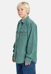 Camicia in corduroy verde con una vestibilità rilassata, fronte con bottoni, due tasche sul petto e una toppa decorativa. Indossata con pantaloni di denim blu.