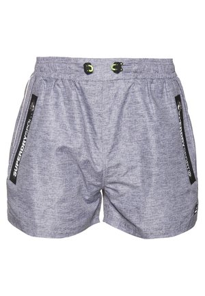 Mężczyzna w szarych sportowych szortach z zamykanymi kieszeniami bocznymi i czarnych butach sportowych, stojący na białym tle.