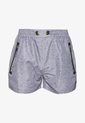 Man draagt grijze sportkorte broek met ritssluitingen aan de zijzakken en zwarte sportschoenen, staat tegen een witte achtergrond.