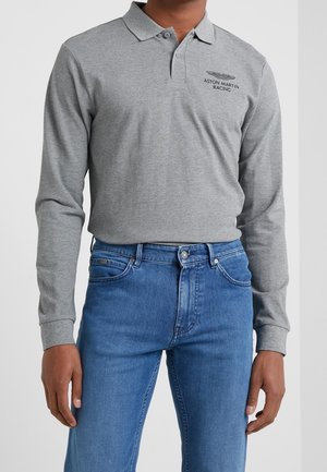 Uomo che indossa una polo grigia a maniche lunghe con il logo Aston Martin Racing e jeans blu, in piedi davanti a uno sfondo uniforme.