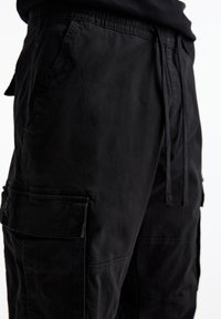 Zwarte cargobroek met een elastische tailleband, trekkoord, zij- en dijzakken, en een gestructureerde stofafwerking.