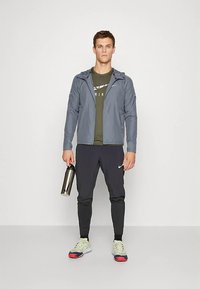 Men's athletic outfit featuring a gray lightweight jacket, green t-shirt, black pants, and sneakers with a bold color sole. Holds a silver water bottle.
