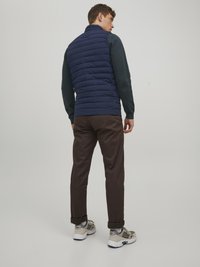 Gilet matelassé bleu marine avec un col haut, associé à un pantalon marron foncé et des chaussures de sport grises, vu de dos contre un fond neutre.