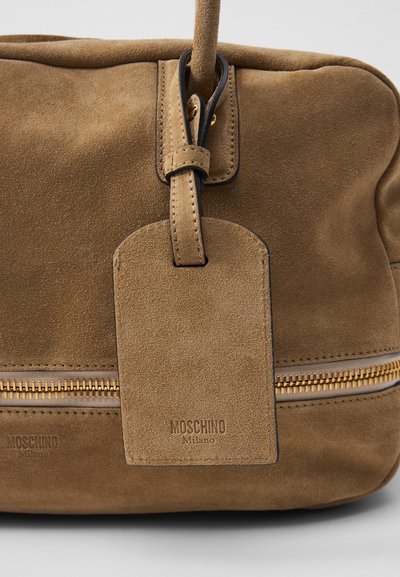 Brązowa zamszowa torba z złotym zamknięciem na zamek, z skórzaną etykietą i wytłoczonym logo. Miękka faktura i zaokrąglony kształt, minimalistyczny design.