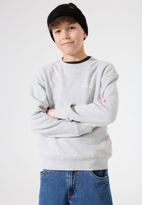 Sudadera gris con puños y bajo acanalados, que presenta un pequeño logo blanco. Combinada con jeans de mezclilla azul y un gorro negro acanalado.
