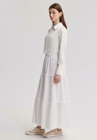 Robe blanche à volants avec un tissu texturé, manches longues et un gilet court. Associée à des baskets beige clair et à des boucles d'oreilles discrètes.