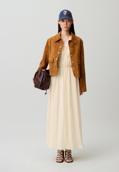 Giovane donna che indossa una giacca in camoscio marrone chiaro, un vestito lungo color crema, un berretto da baseball blu navy, tiene in mano una borsa marrone in pelle con coulisse e indossa sandali neri con cinturini.