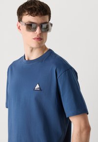 Blå bomulds t-shirt med korte ærmer og rund halsudskæring, med et trekantet broderet logo på brystet. Stoffet har en glat overflade.