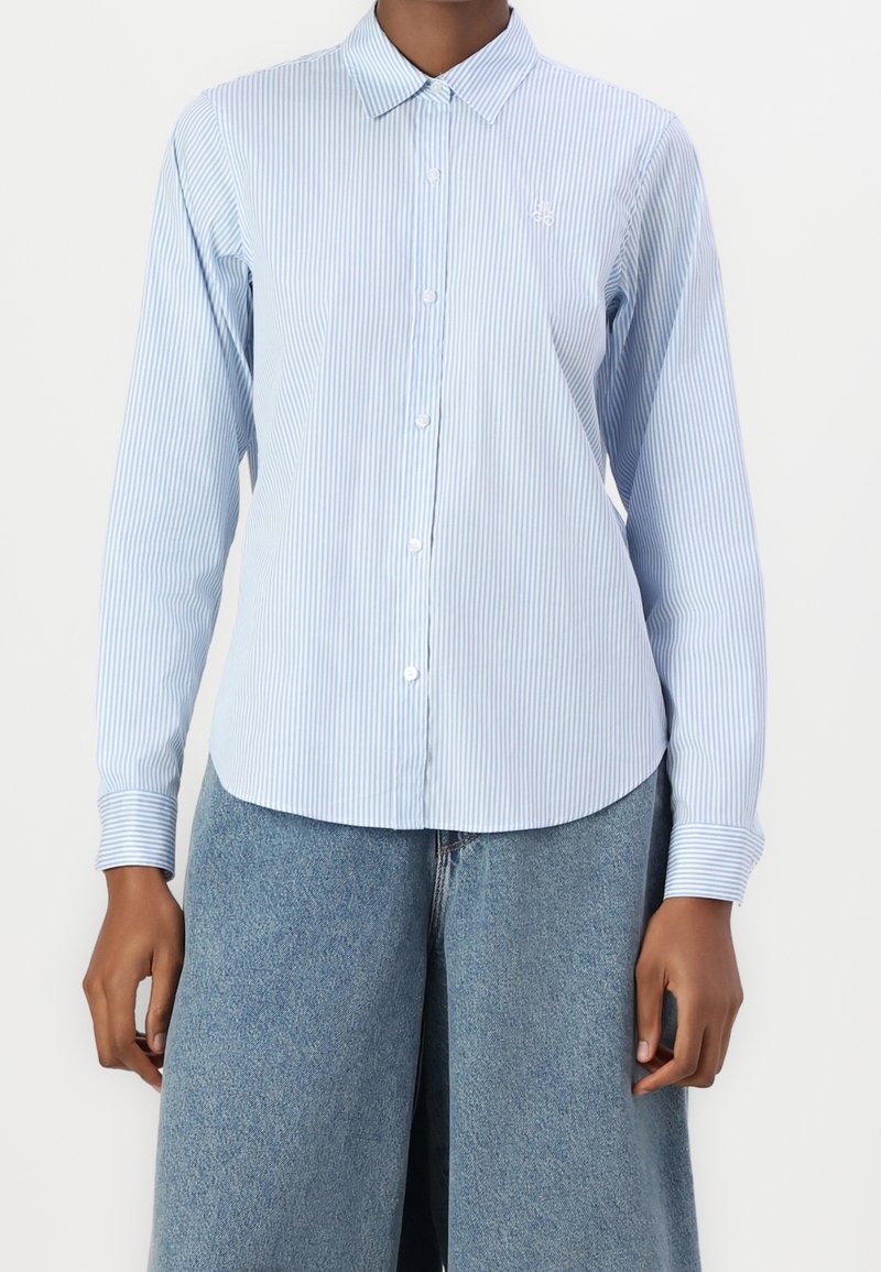 Femme portant une chemise boutonnée à fines rayures verticales bleu clair et blanches avec des manches longues et un jean en denim bleu.