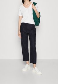 Vit t-shirt med v-ringad hals som matchas med marinblå kapade byxor med en subtil textur. Vita sneakers och en grön jacka hängande över en axel.