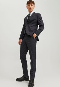 Completo su misura grigio scuro con giacca a un bottone, rever a punta e pantaloni slim-fit. Indossato con una camicia bianca e una cravatta nera. Scarpe eleganti nere.